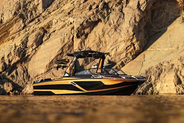 A black and yellow wakeboat in the water near a rocky cliff.