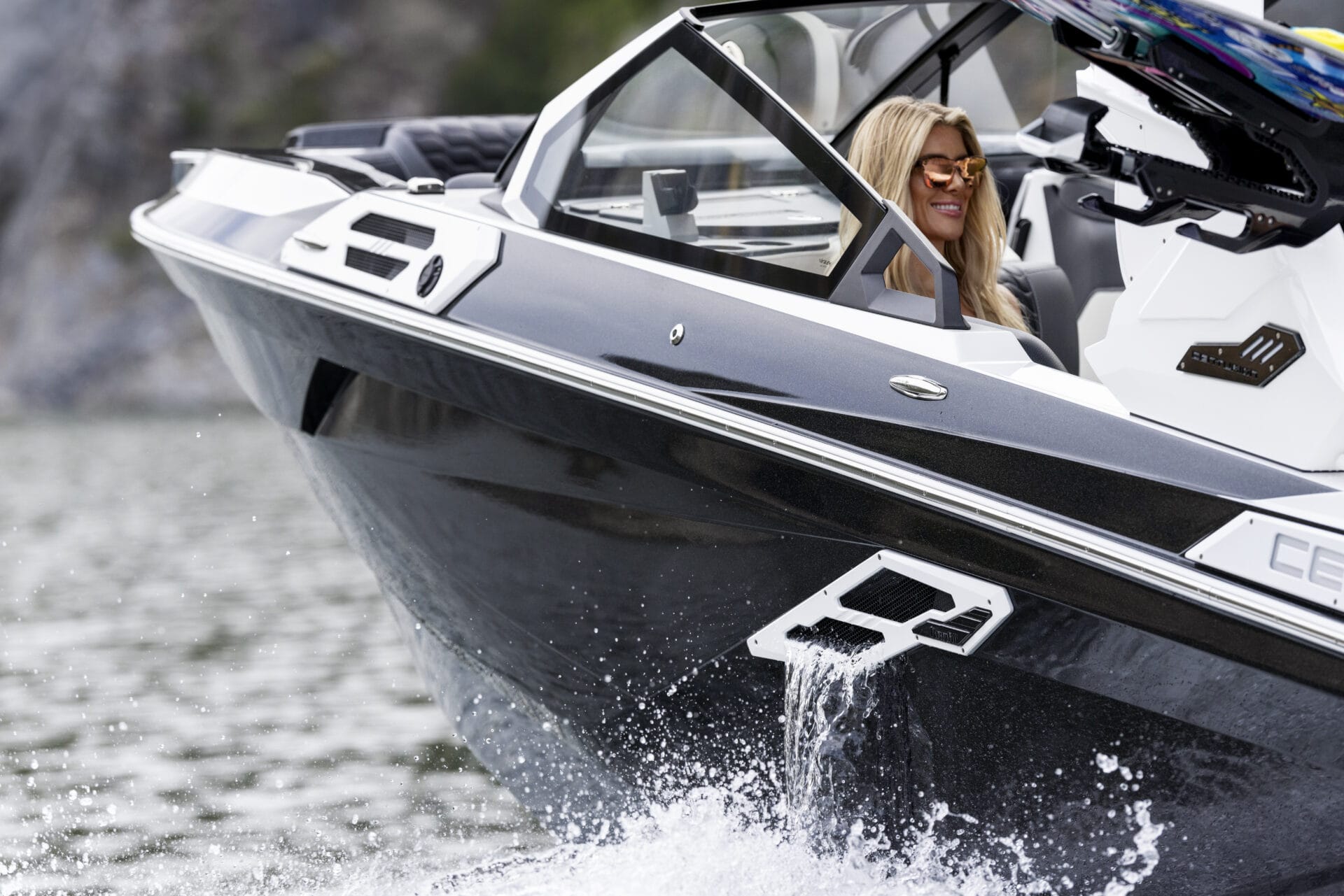 A woman in sunglasses is smiling while seated at the steering wheel of a sleek, black speedboat on the water, embodying the spirit of Customize My Ride.