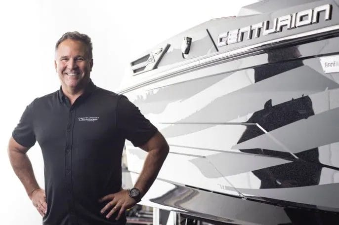 A man posing with a wakesurf board and a wakeboat.