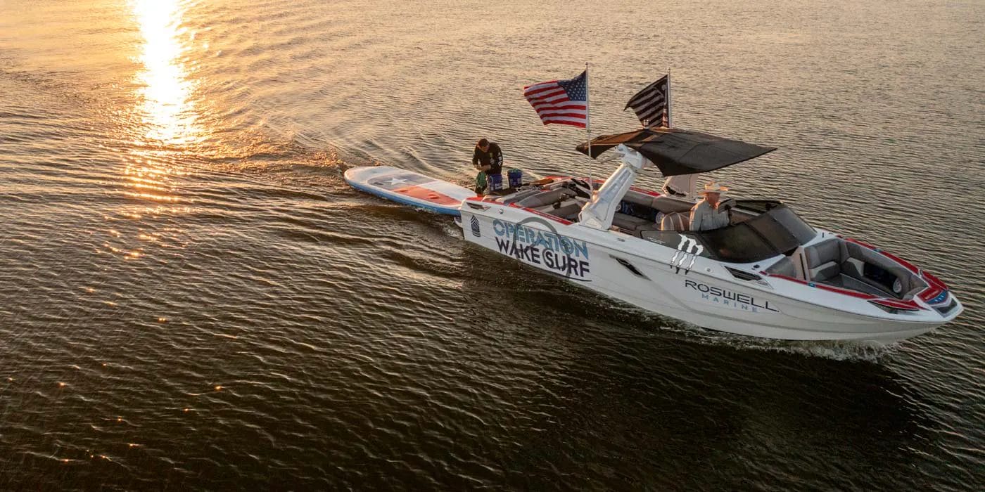 A group riding a wakesurf boat in the water at sunset.