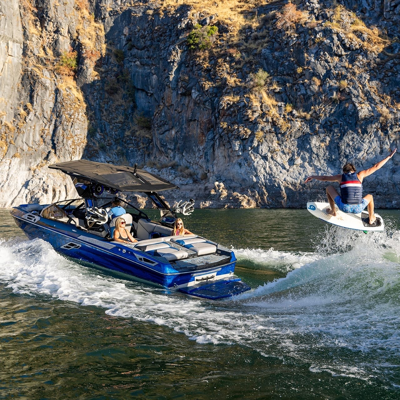 A person wakeboards behind a blue Centurion Ri230 motorboat on a lake, surrounded by rocky cliffs.