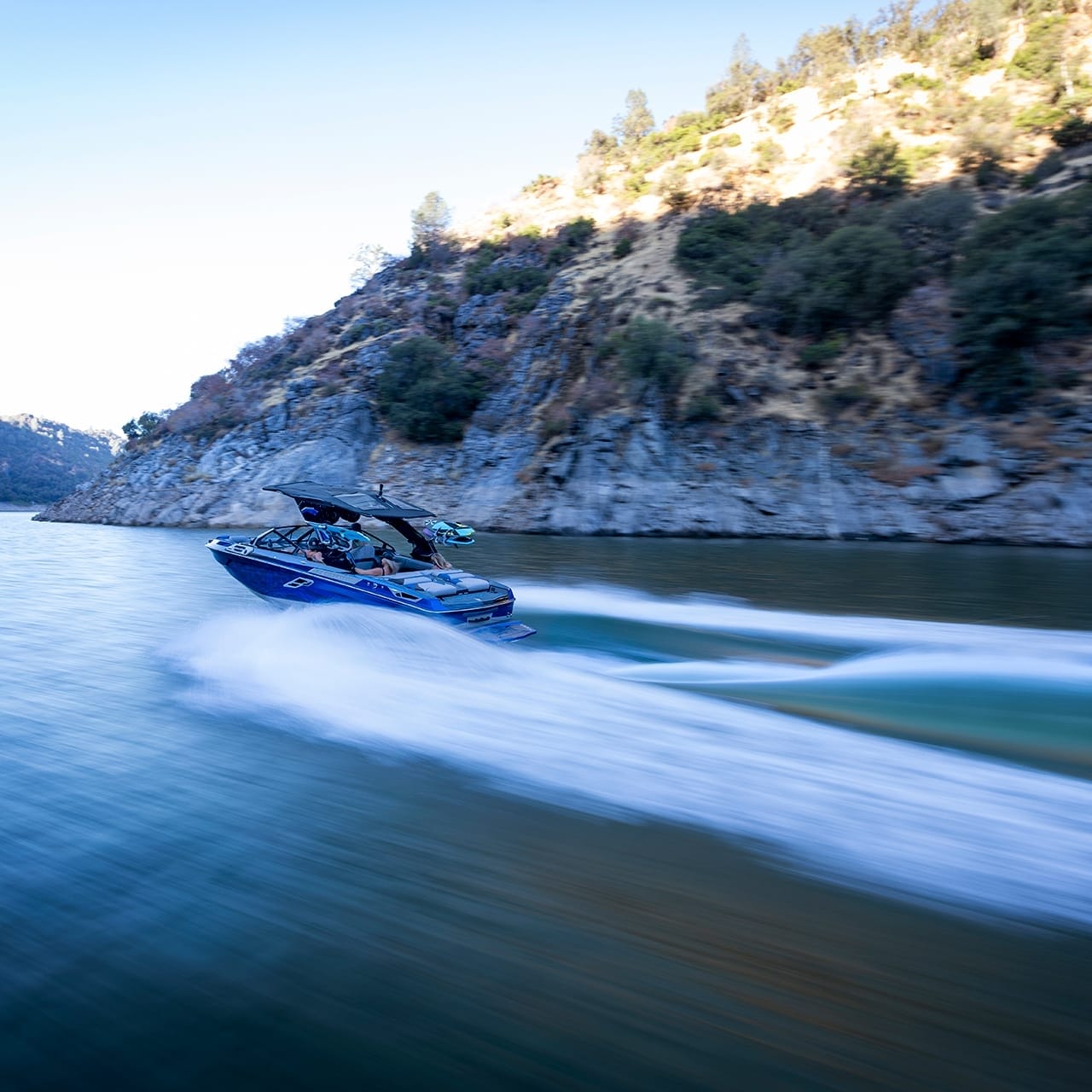 The Centurion Ri230 motorboat speeds through a narrow, winding waterway with rocky hills and sparse trees on both sides, creating wakes on the water.