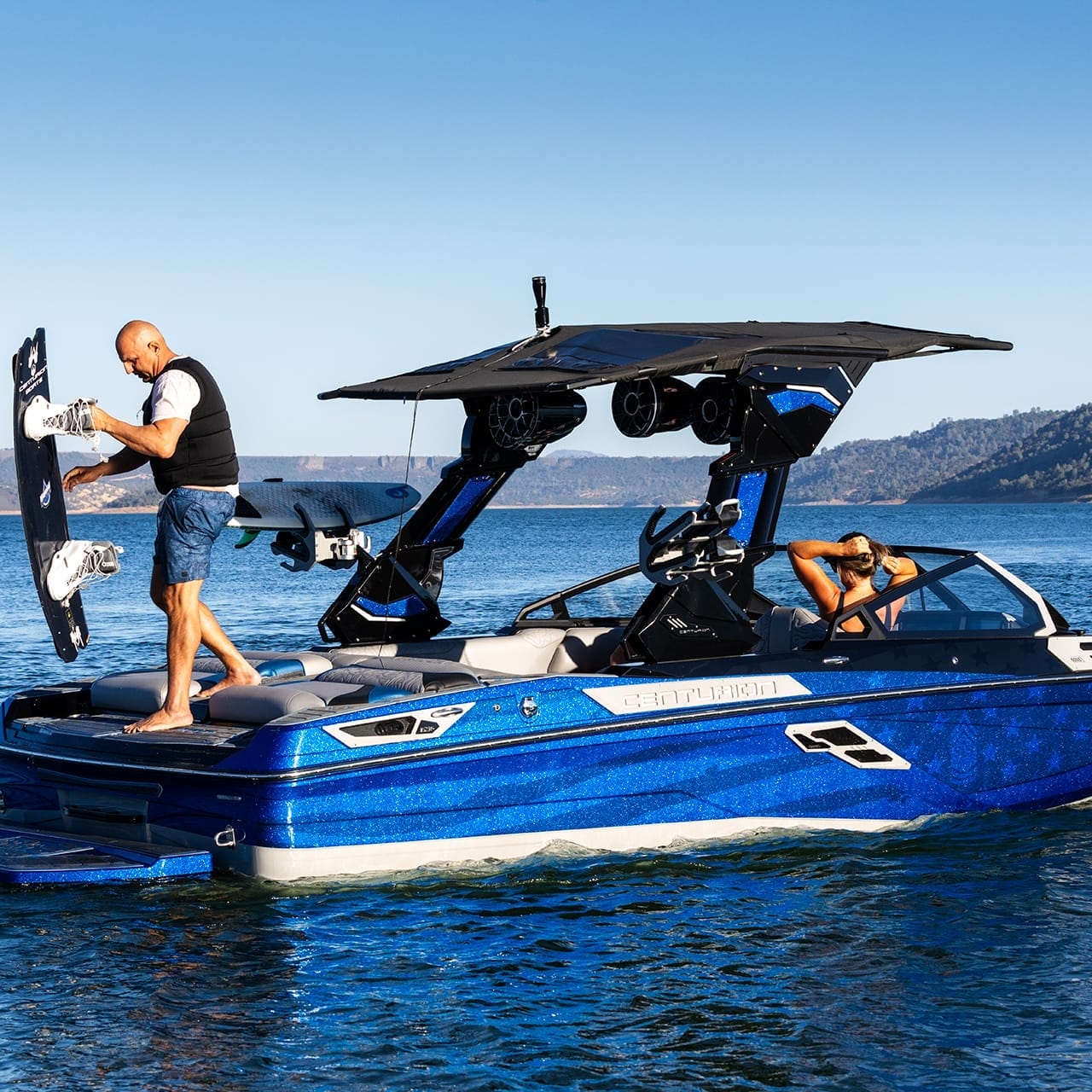 A man prepares to wakeboard from a sleek Centurion Ri230 boat with a tower, gliding on a calm lake under the clear blue sky.