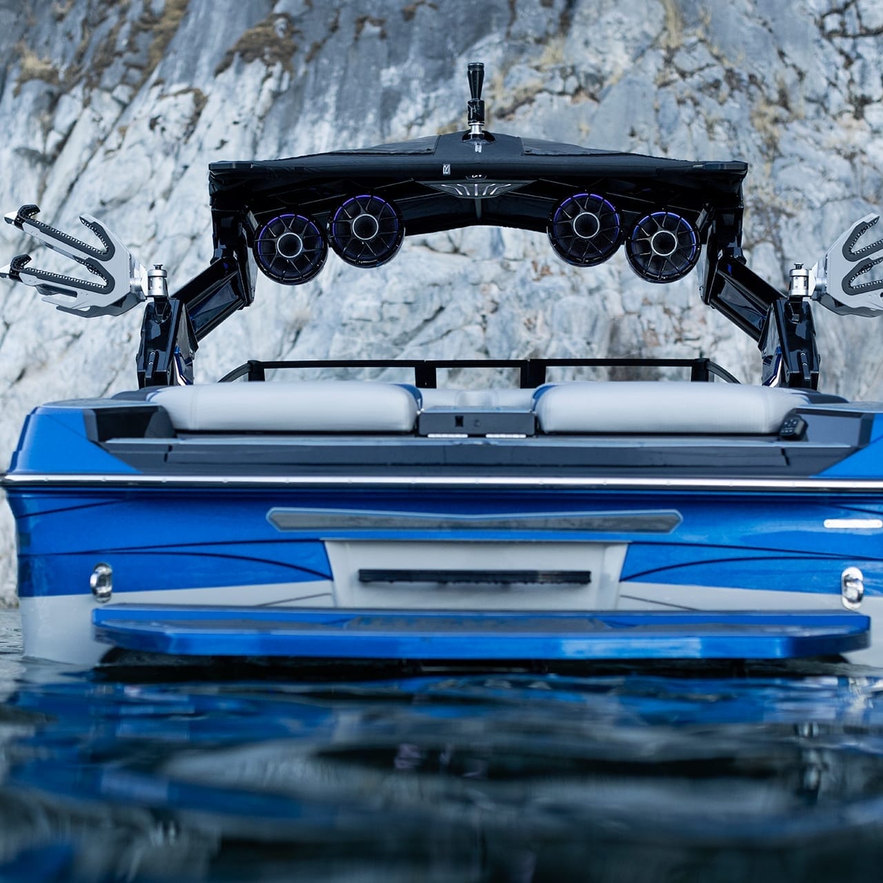 A blue Centurion Ri230 boat with speakers and racks on the deck glides effortlessly on the water in front of a rocky cliff.