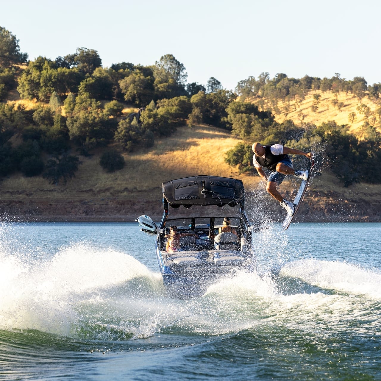 A person wakeboarding behind a Centurion Ri230 performs an airborne trick on a lake, with hilly terrain and trees in the background.