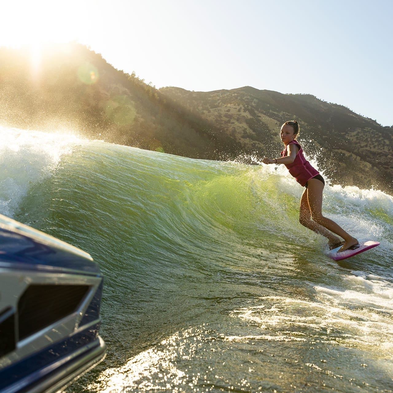 A person in a swimsuit skillfully surfs a wave created by the sleek Centurion Ri230 on a sunny day, with hills painting the serene background.