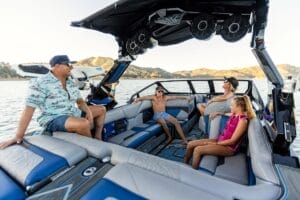 Four people relax on a Centurion Ri230 boat on a lake, surrounded by mountains. The boat boasts blue and gray seating and large speakers, perfect for enjoying the serene landscape.