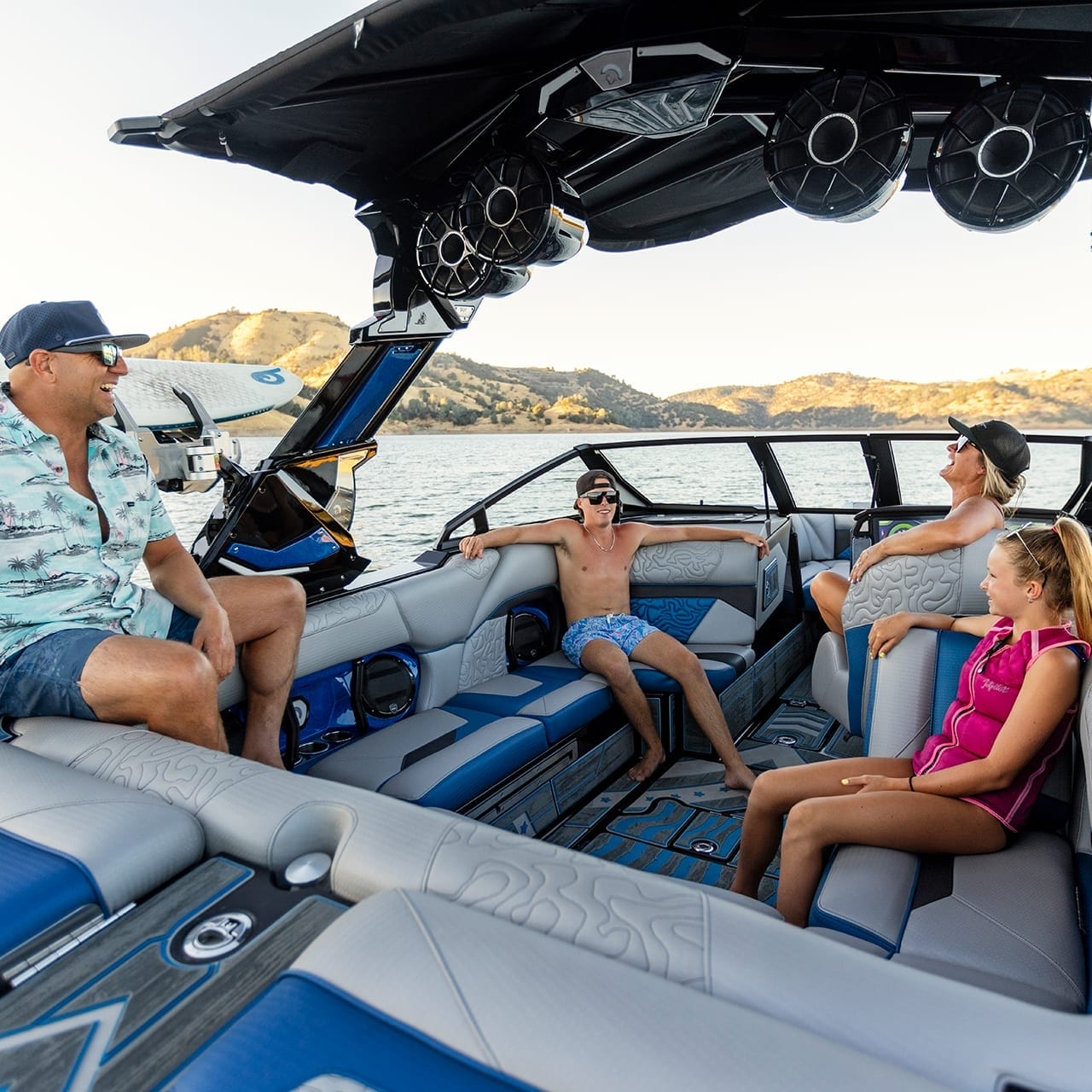 Four people relax on a Centurion Ri230 boat on a lake, surrounded by mountains. The boat boasts blue and gray seating and large speakers, perfect for enjoying the serene landscape.