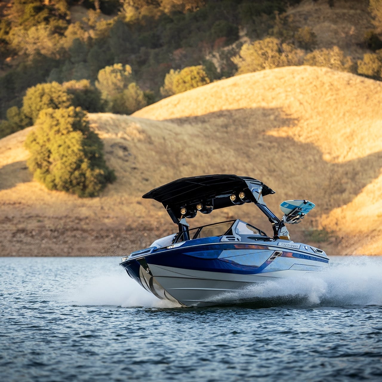 A Centurion Ri230 with a black canopy speeds across the lake, its sleek blue design contrasting against the backdrop of hills and trees.