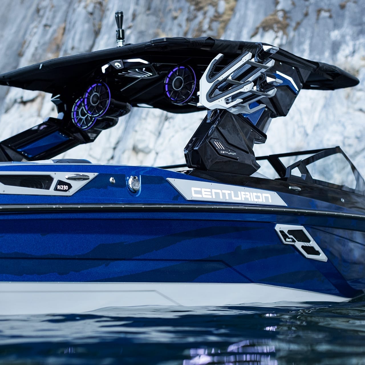 A blue Centurion Ri230 with large speakers is docked near a rocky shore.