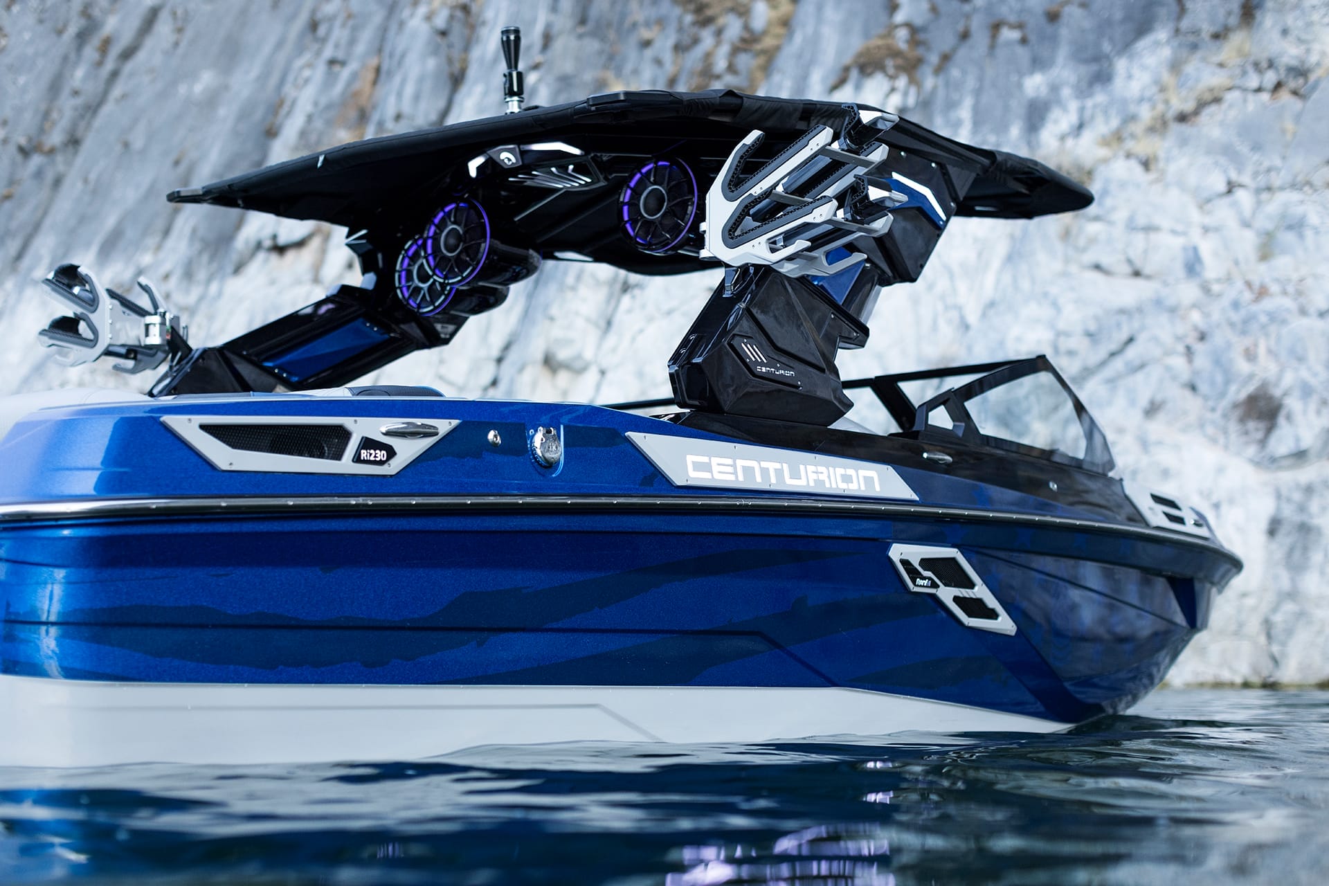 A blue Centurion Ri230 with large speakers is docked near a rocky shore.