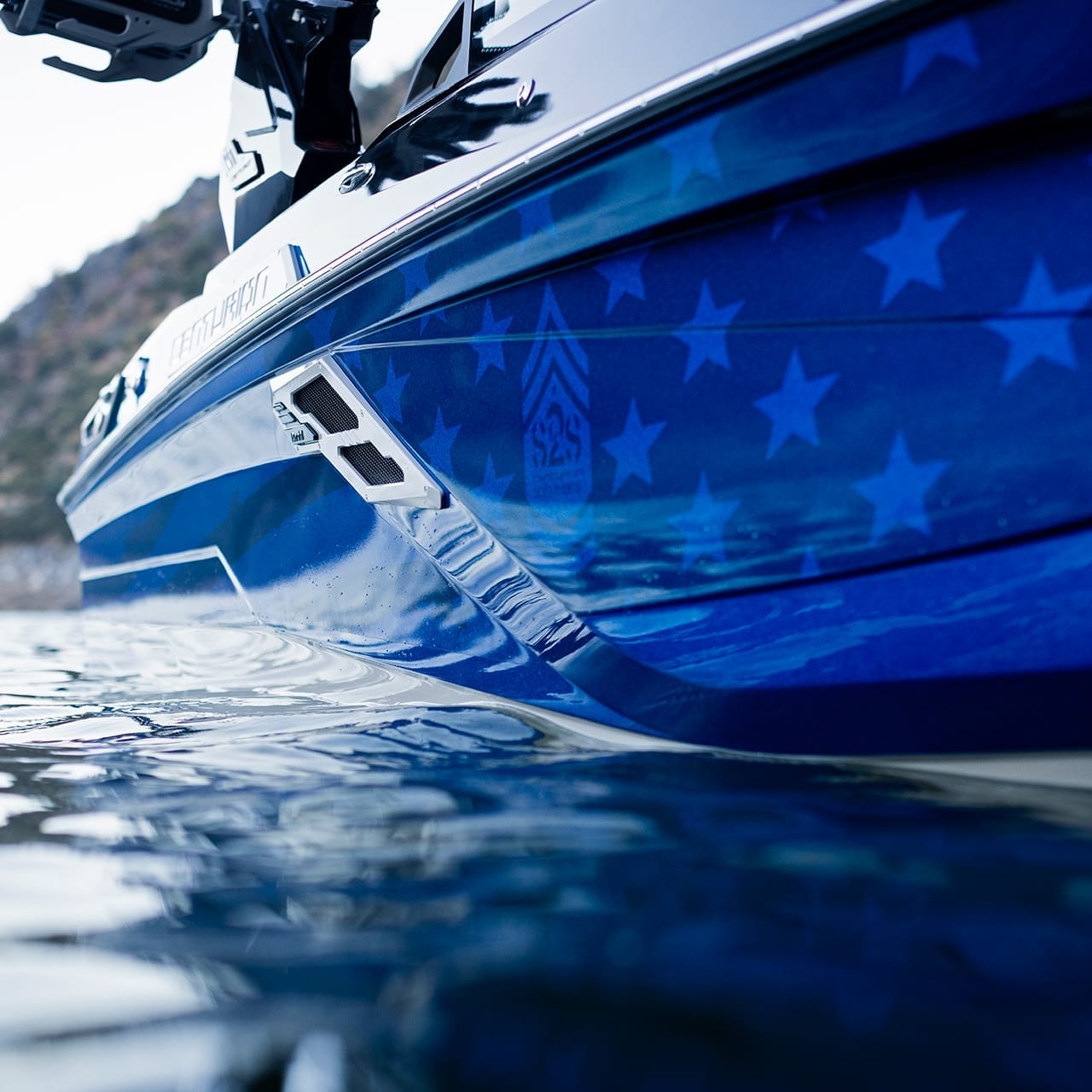 A close-up of a blue Centurion Ri230 boat with star patterns graces the tranquil water, its sleek form mirrored on the surface while a distant shoreline lingers in the background.