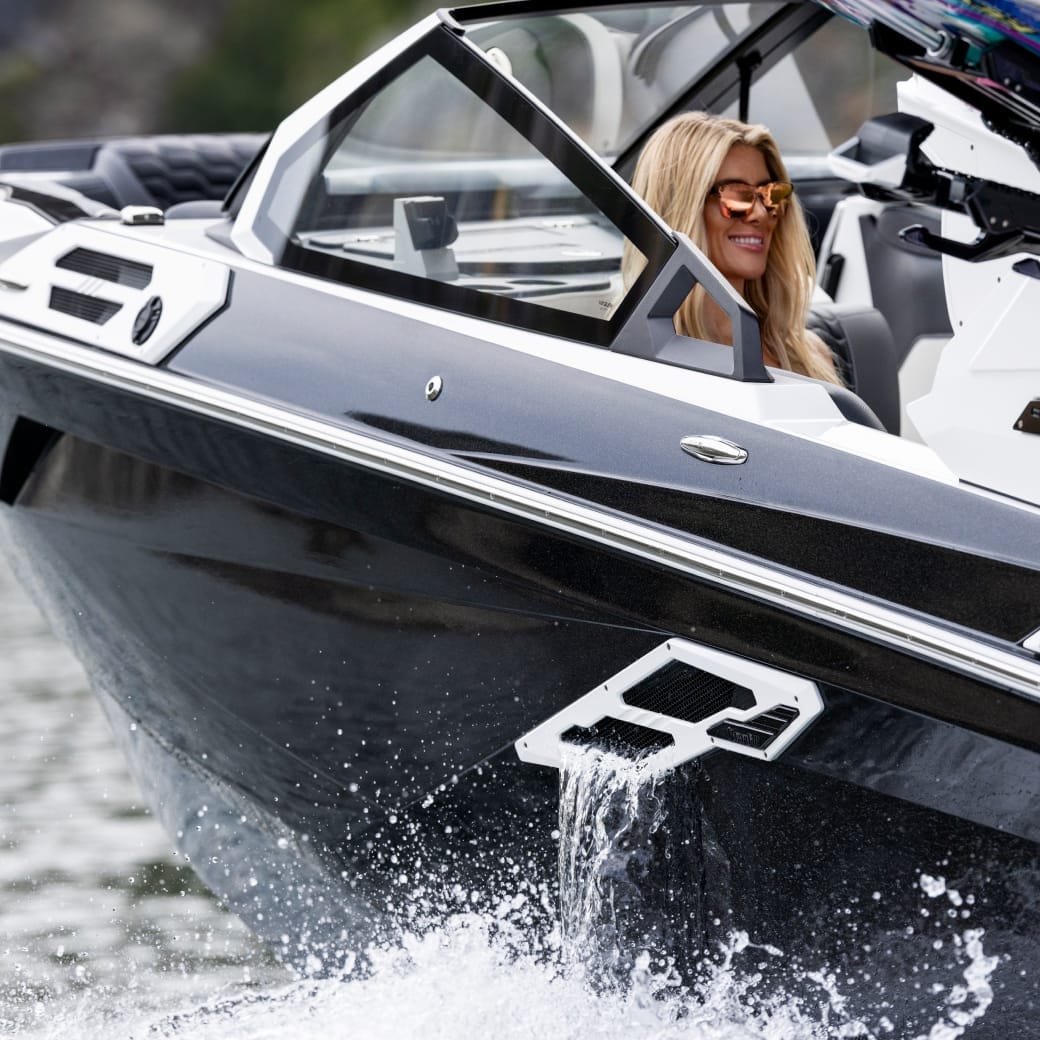 A person with long blond hair and sunglasses is driving a Centurion Ri245 speedboat on the water, which is creating a splash.