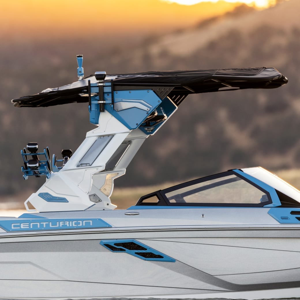 A blue and white Centurion Ri245 speedboat with a unique tower featuring wakeboard racks is set against a blurred background of sunlight and hills.