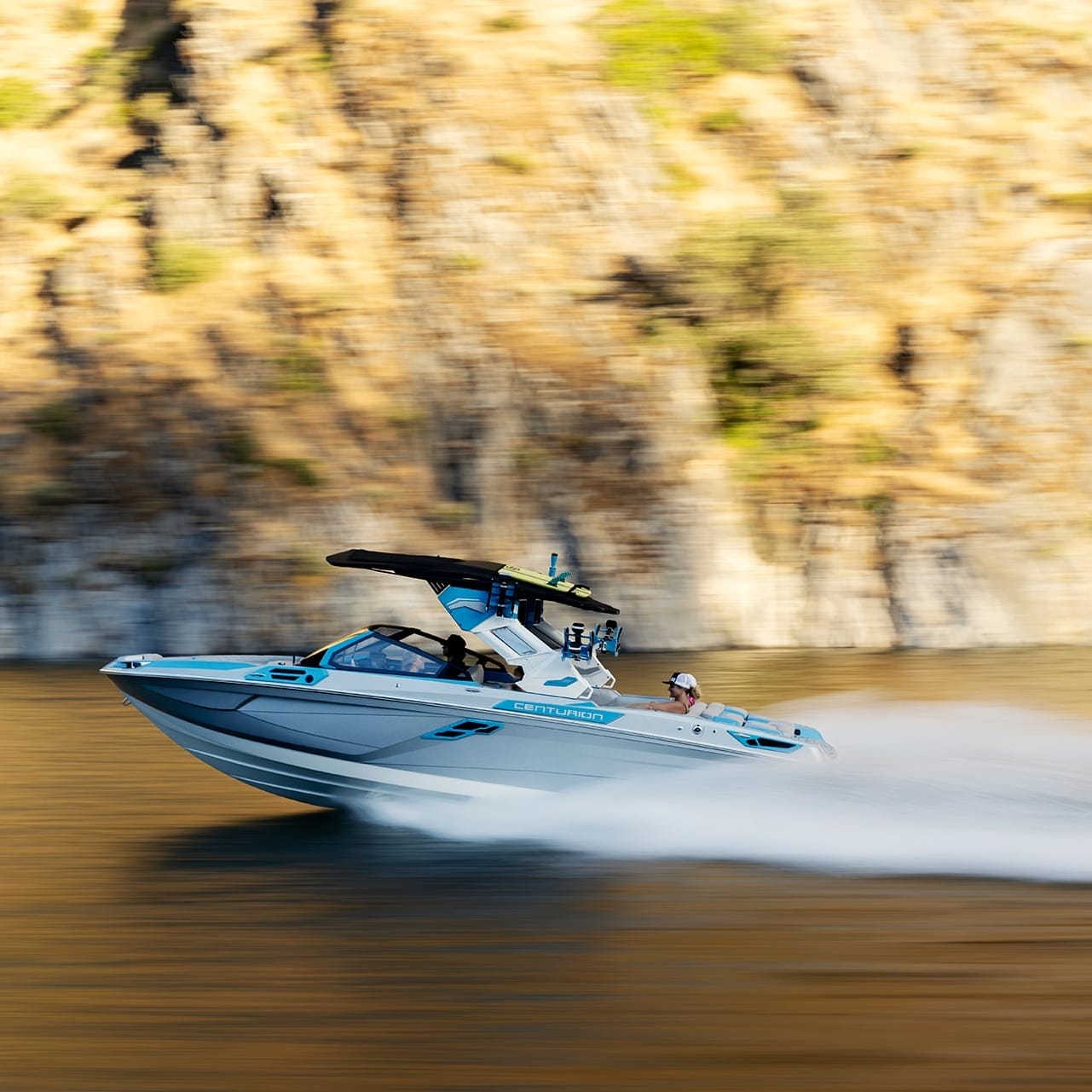 A Centurion Ri245 speedboat glides quickly across a calm body of water with a rocky, sunlit shoreline in the background.