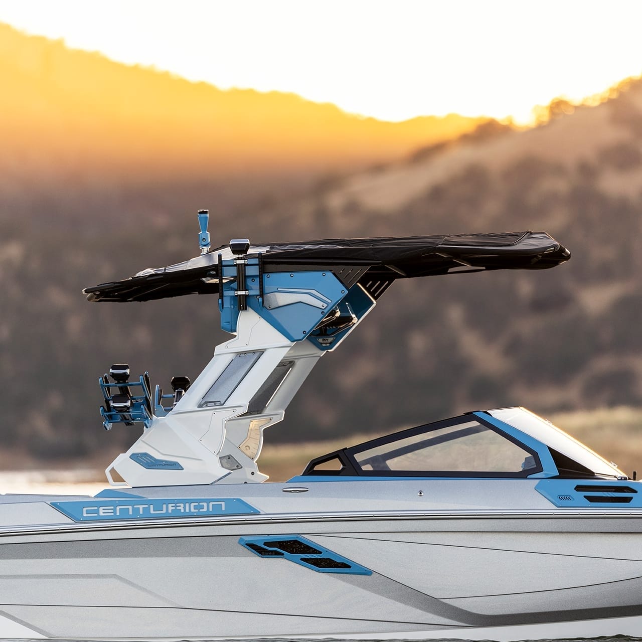 A blue and white Centurion Ri245 boat on water with a wakeboard tower, captured against a backdrop of hills and a golden sunset.