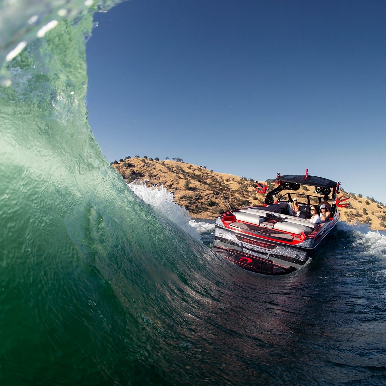 A Centurion Ri265 speedboat with people on board navigates on a clear blue water body under a sunny sky, creating a large wave beside the boat. Hills are visible in the background.