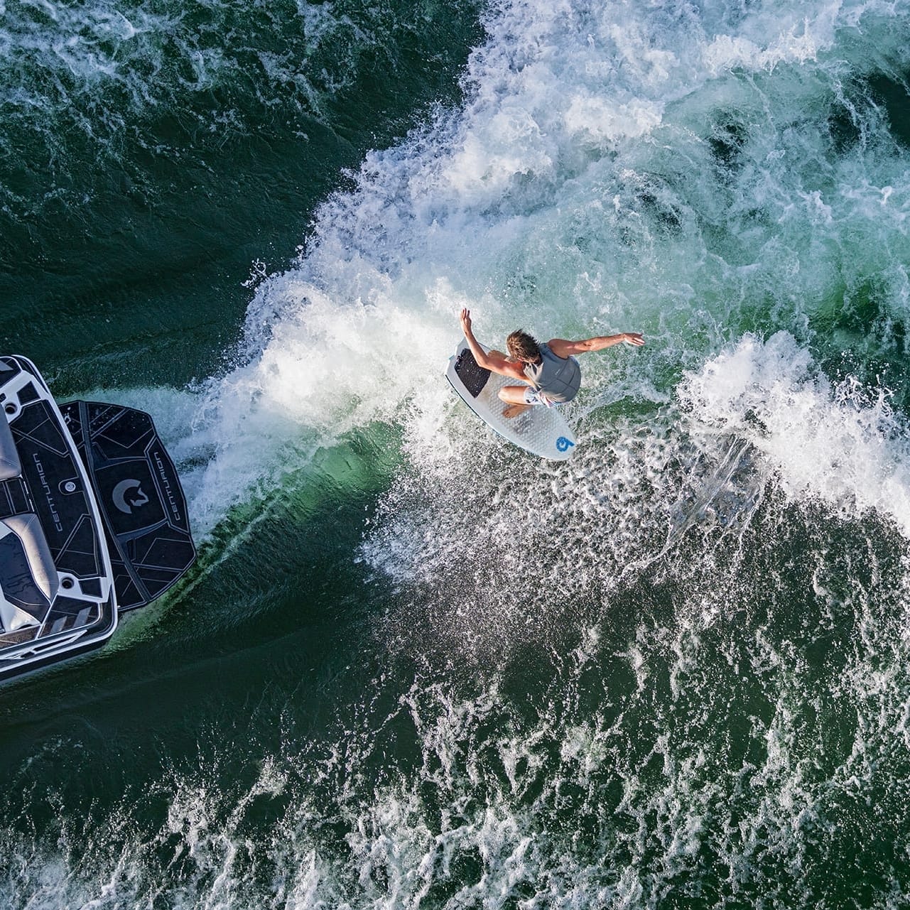 A person rides a surfboard through a boat's wake on a body of water, catching thrilling waves created by the powerful Centurion Ri265, while another person observes from the boat.