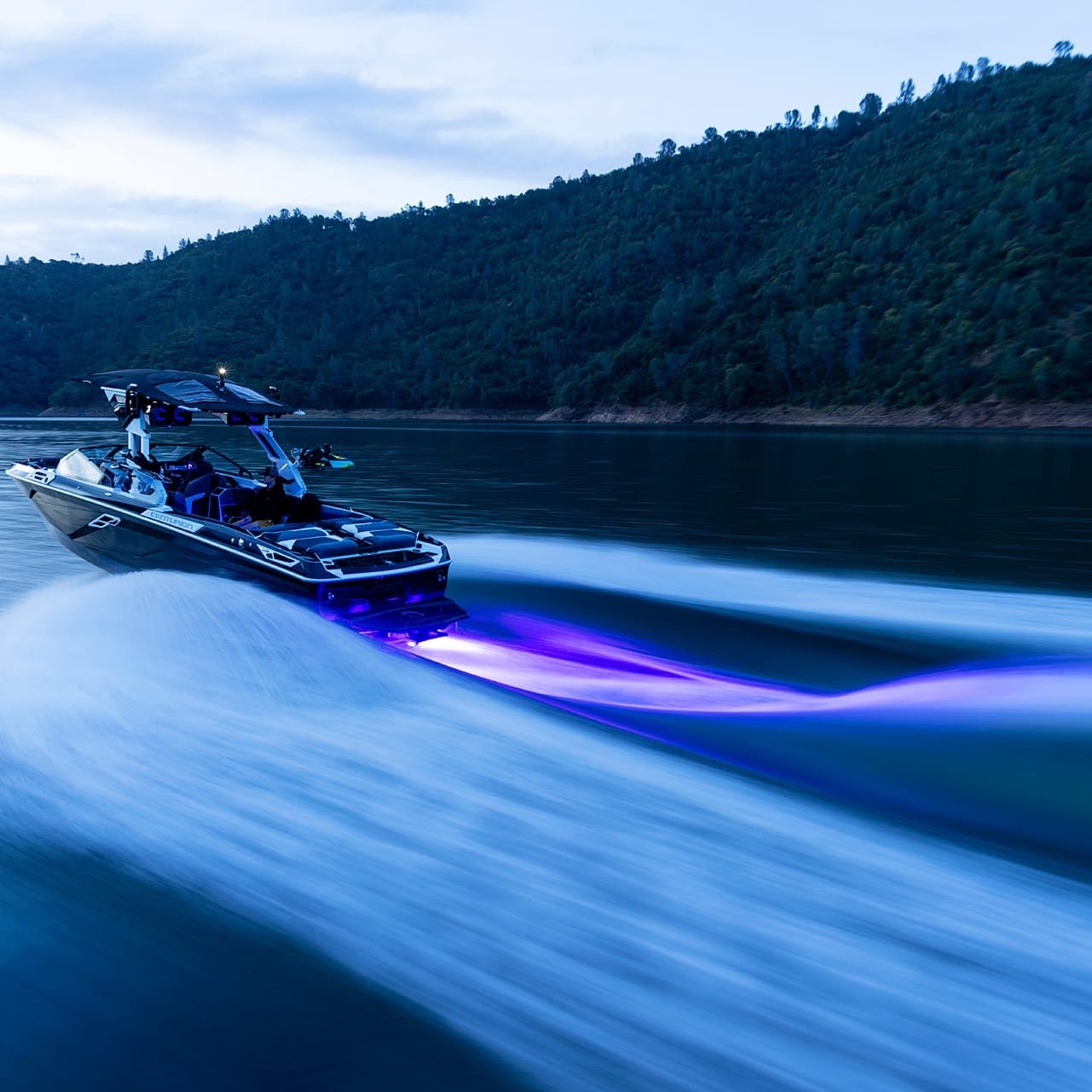 The Centurion Ri265 speedboat, adorned with blue lights, travels swiftly on a lake, creating waves. Mountains and trees are visible in the background under a cloudy sky.