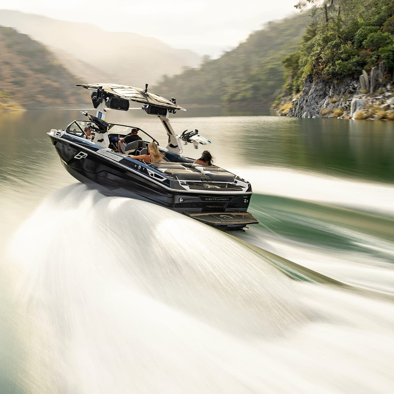 A black Centurion Ri265 motorboat speeds across a serene, narrow river flanked by forested hills, creating white wake trails.