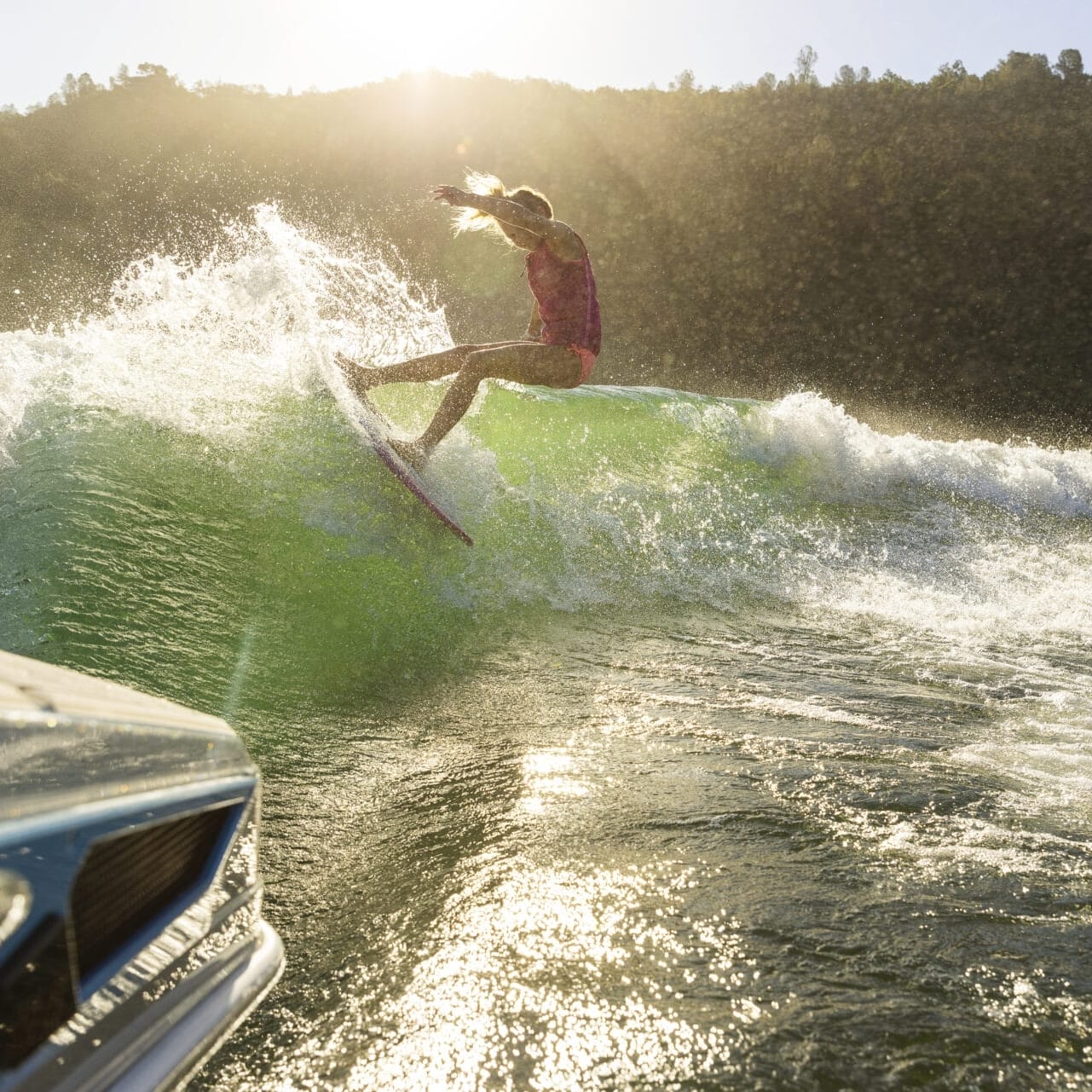 Kenzie Hickey surfs gracefully on a wave created by a boat, silhouetted against the golden sunlight in the background.