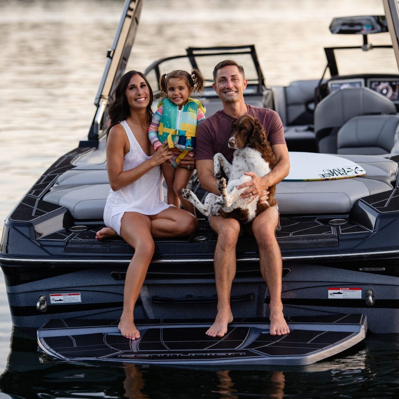 A family of three and a dog sit together on the back of a boat on a calm body of water.