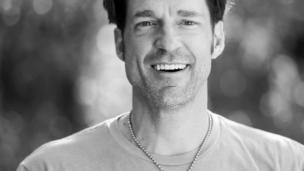 A black and white photo of a man smiling while wakesurfing behind a wakeboat.