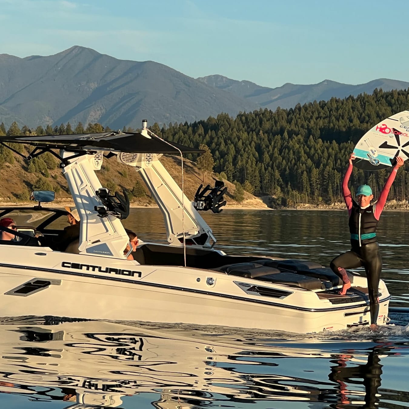 A man is standing on a wakeboat with a wakesurf board in the water.