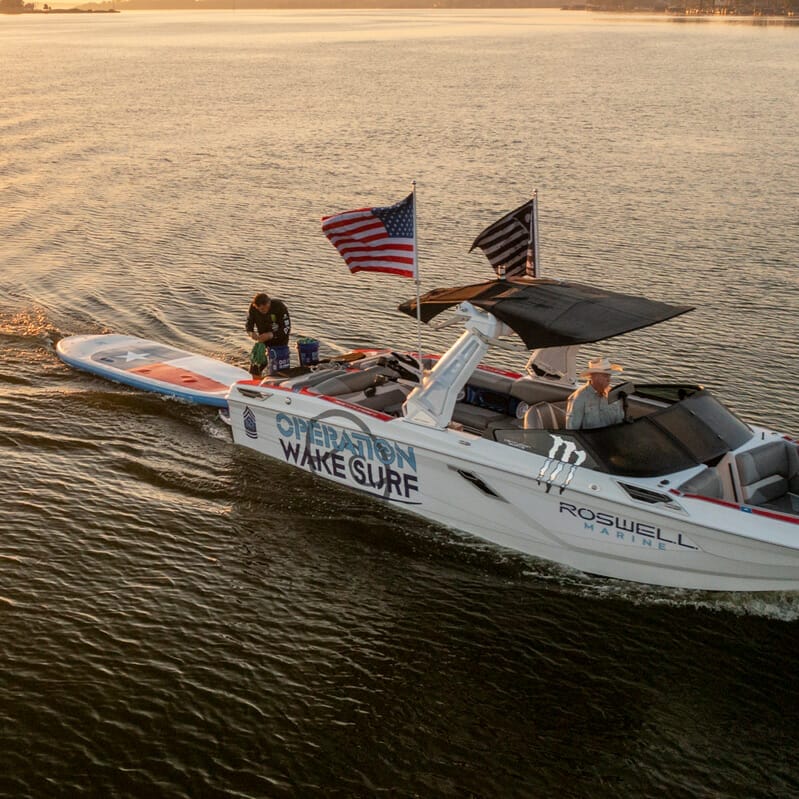 A wakesurf boat with two people on it at sunset.