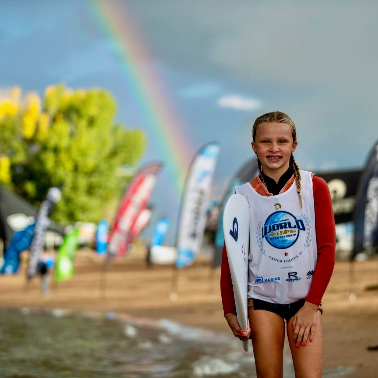 A young person in a wetsuit and jersey, likely Kenzie Hickey, stands on a sandy beach holding a board. Colorful flags flutter in the background near the water with a vibrant rainbow gracing the sky.