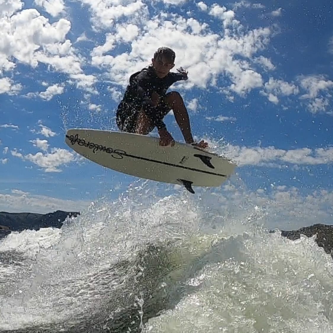 A man riding a wakesurf board in the air behind a wakeboat.