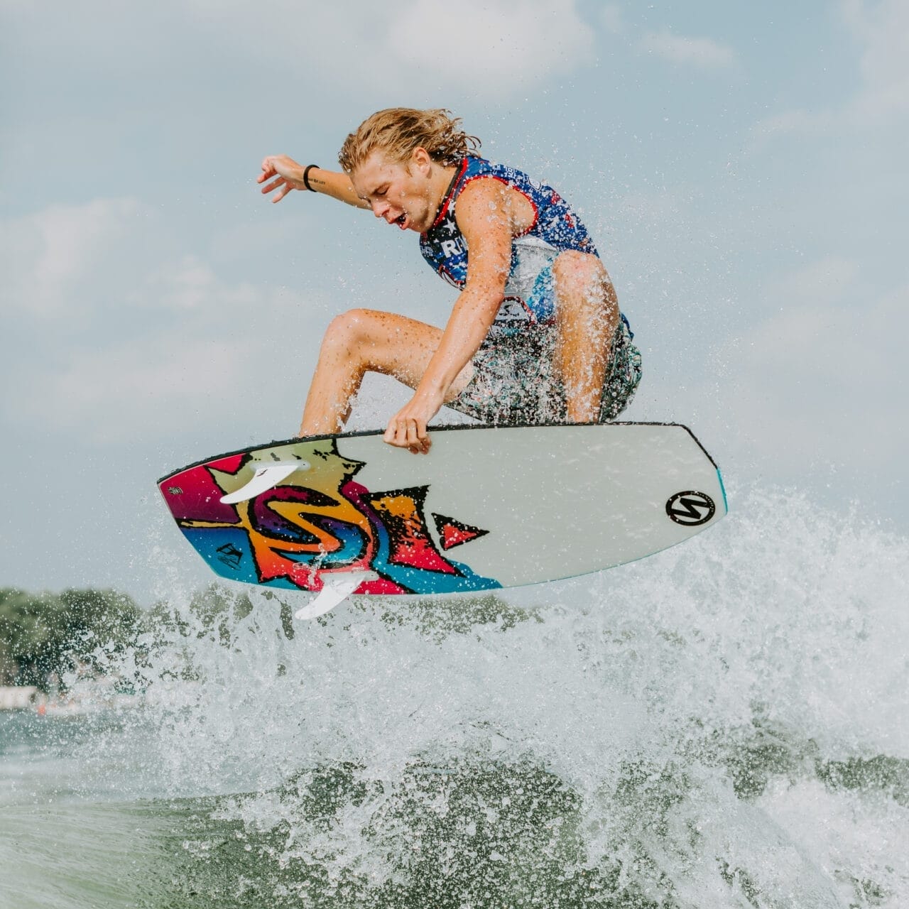 Person performing an aerial trick on a colorful surfboard above a wave, with a partly cloudy sky in the background.