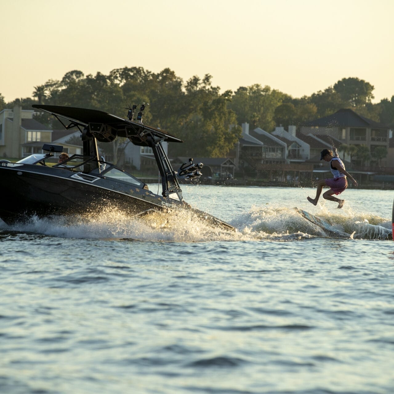 A person is wakeboarding behind a black boat on a lake, with houses and trees in the background and a large red buoy in the water nearby.