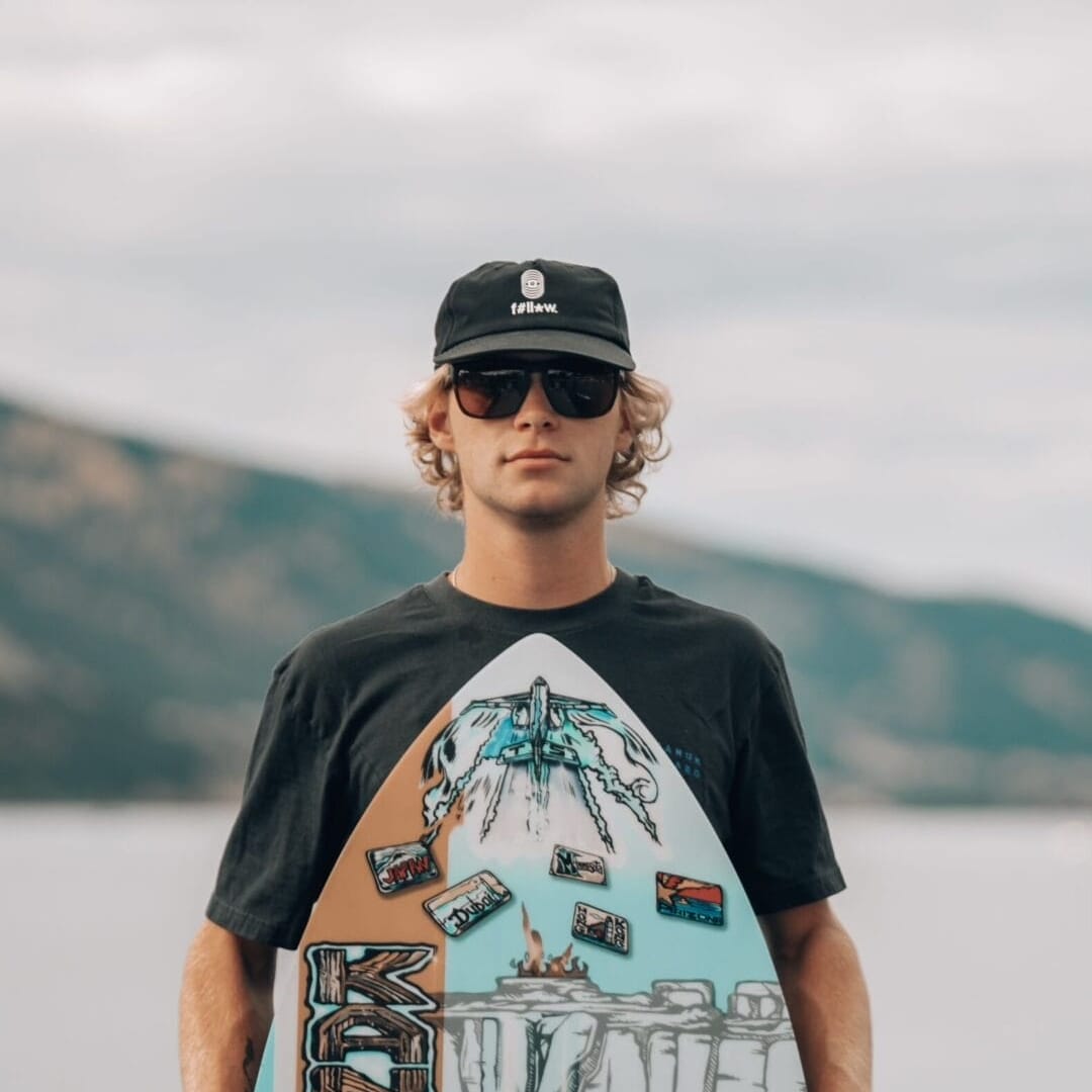 Person wearing sunglasses and a cap, standing in front of water, holding a decorated skimboard.