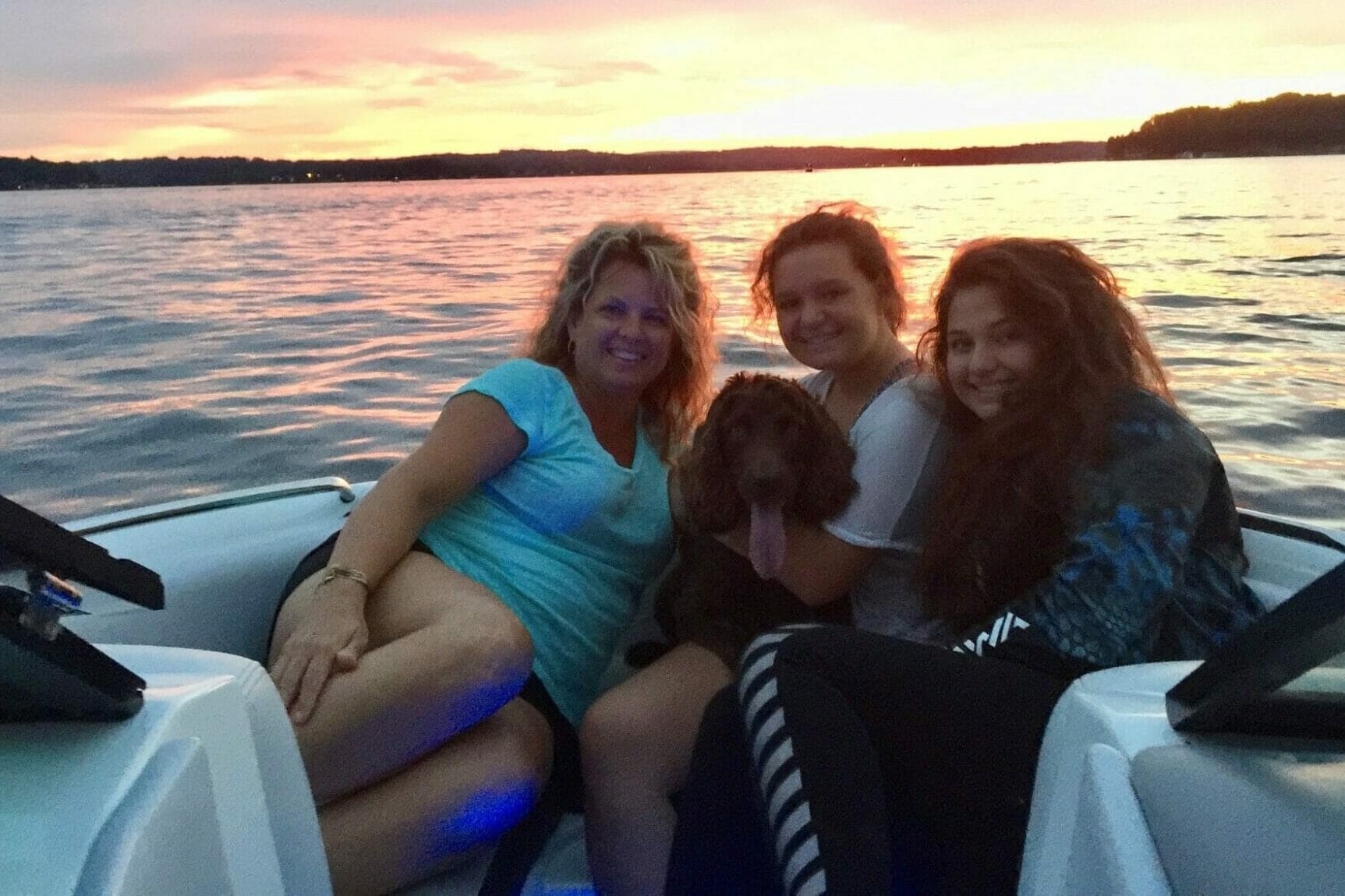 Three women and a dog on a wakesurf boat at sunset.