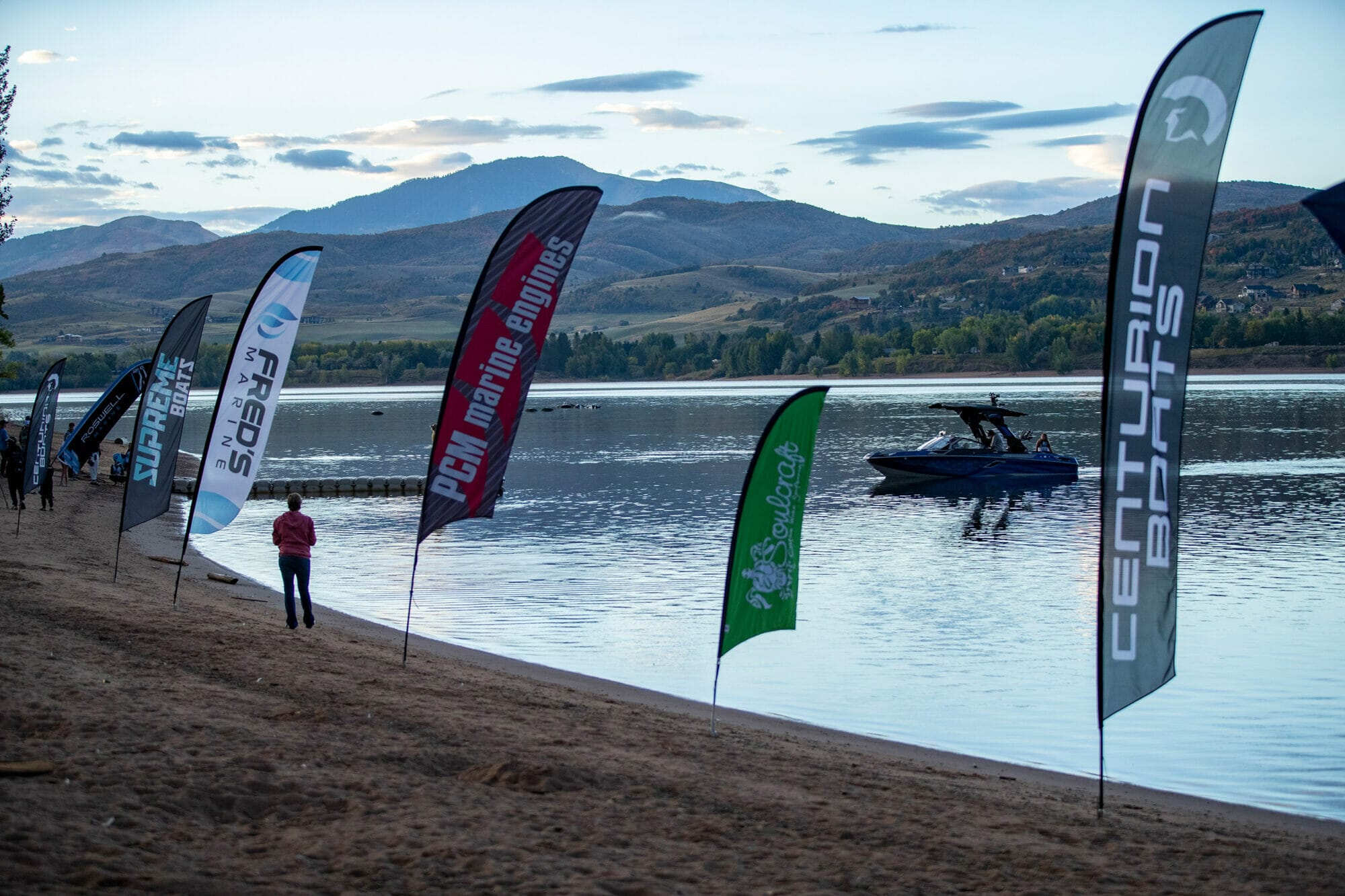 A group of flags on a beach near a body of water, wakesurf boat.