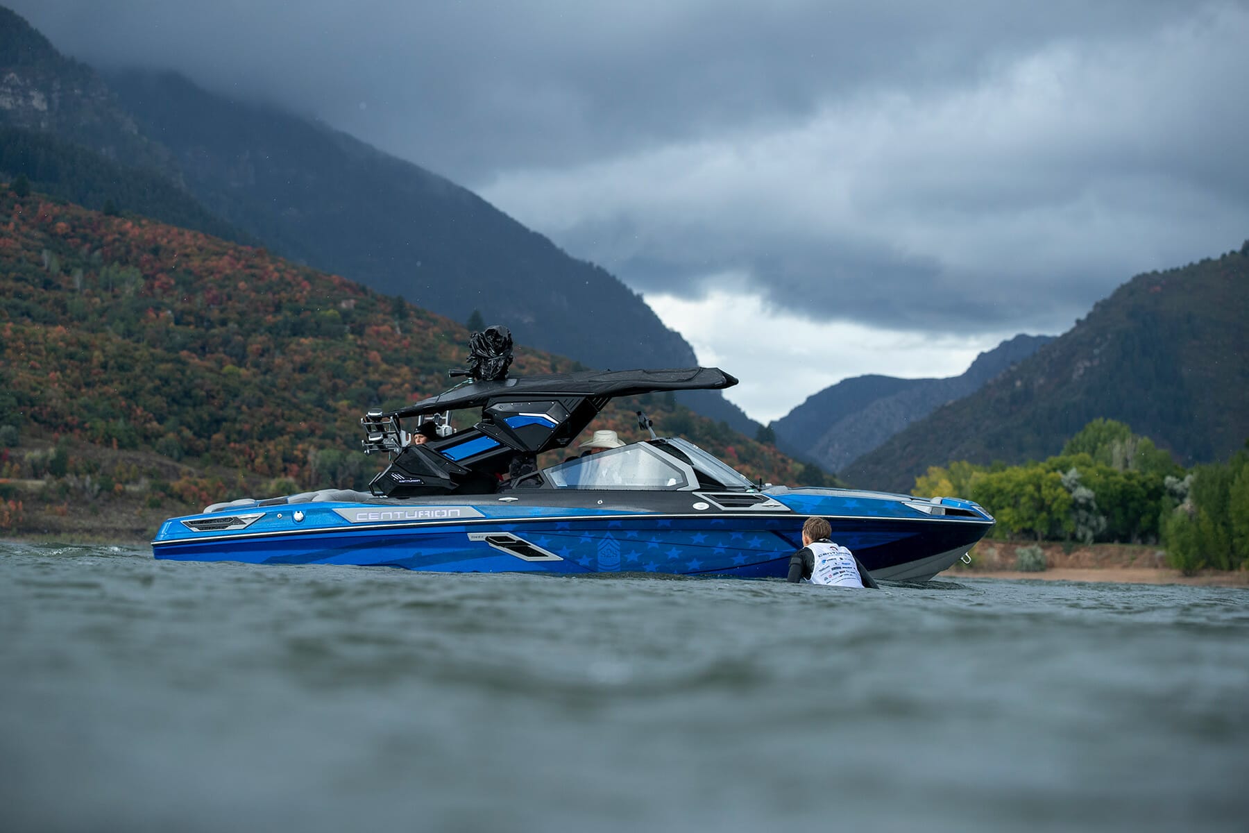 A man in a blue boat on the water with mountains in the background.