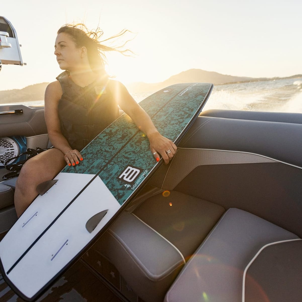 A person in a life vest holds a surfboard while sitting on a Centurion Fe22 speedboat with a scenic backdrop of water and hills during sunset.