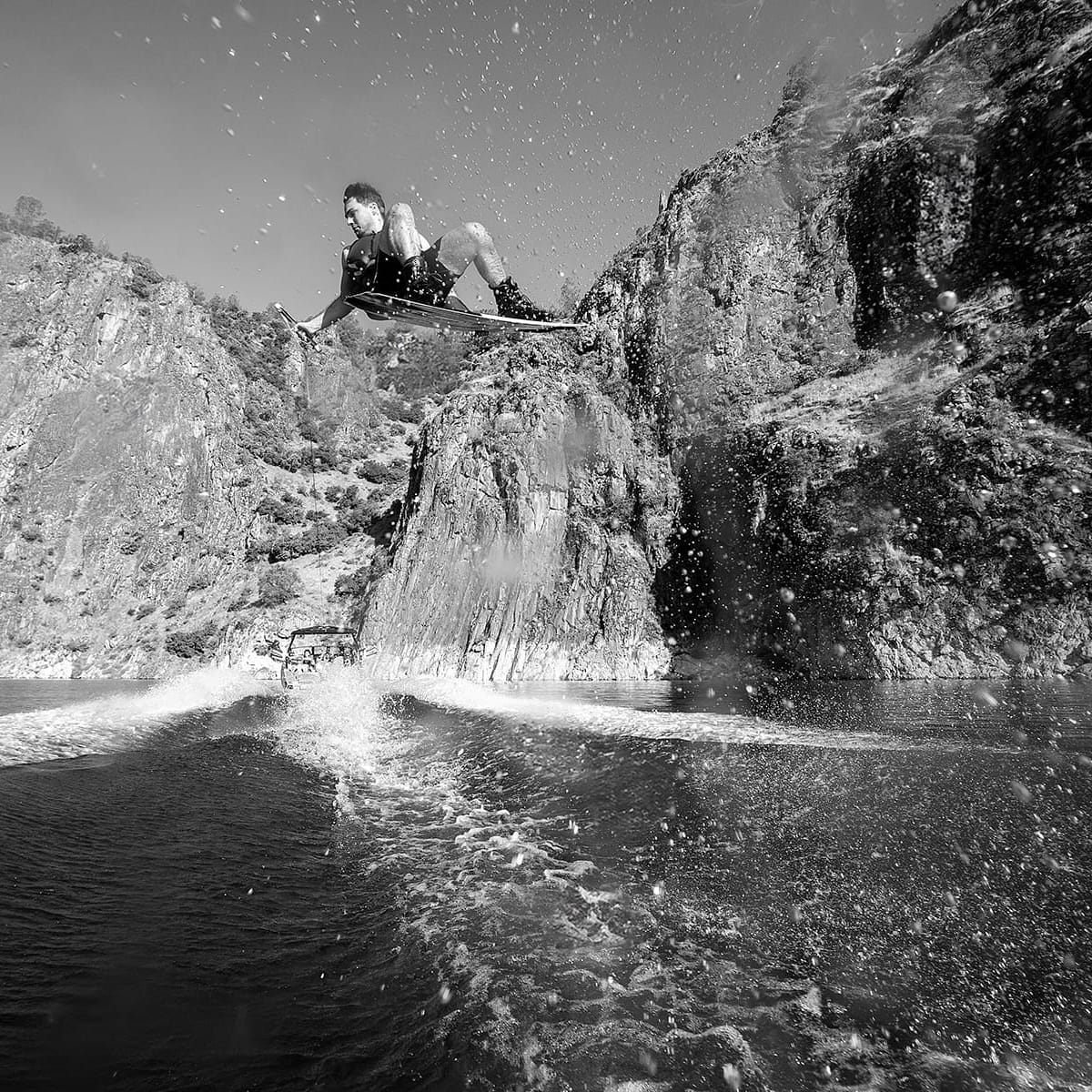 A person performs an aerial trick on a wakeboard above a body of water, with rocky cliffs in the background and water splashing around, all while riding behind a sleek Centurion Fe22.