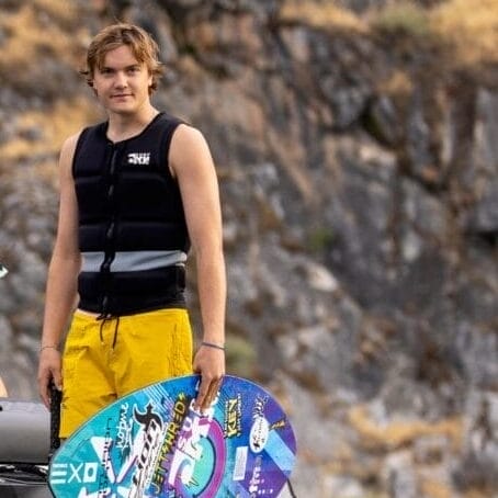 Cade Lybeck stands confidently on a wakeboard beside the boat gliding over calm water, while two others relax inside. Rocky cliffs tower in the background, framing the serene scene.