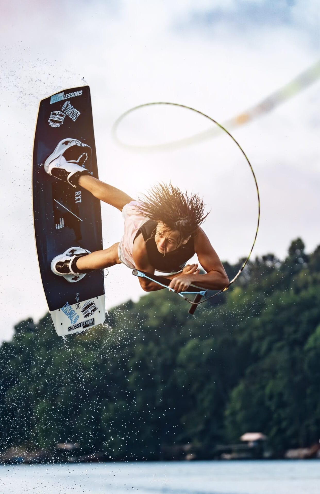 A wakeboarder performs a mid-air trick with a rope above a lake, creating a curved splash of water against a cloudy sky.