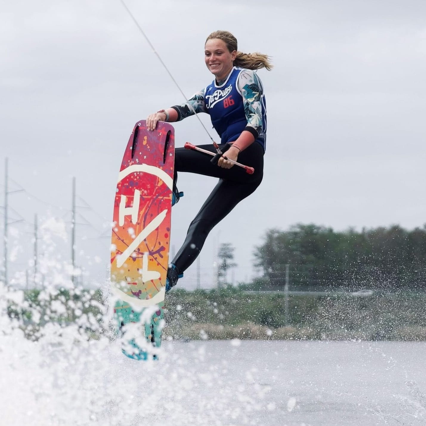 Kitt Smith is wakeboarding in midair over water, holding the handle of a tow rope, with a colorful wakeboard and a slightly cloudy sky in the background.