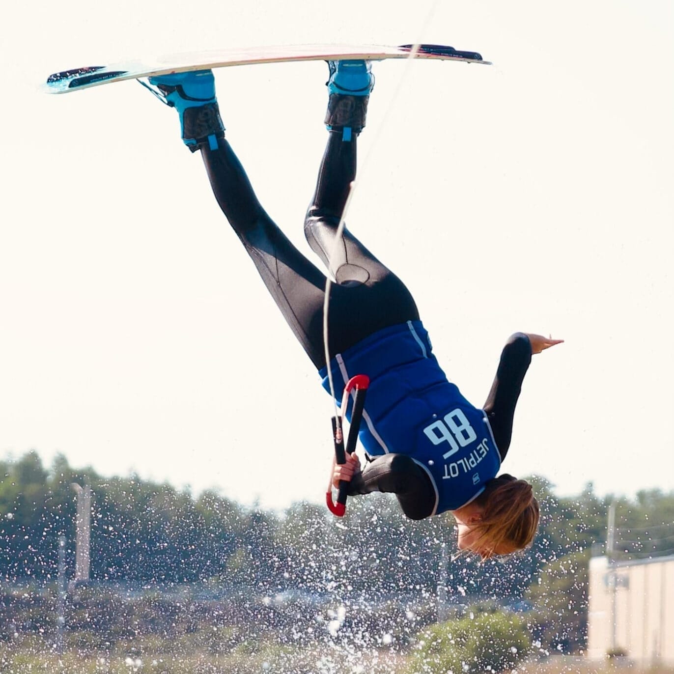 Wakeboarder performing an inverted trick over water, holding the board above with a cable attached, wearing a blue wetsuit and helmet.