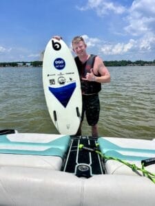 A man stands in shallow water near a boat, holding a surfboard with various logos. He is smiling and gesturing with his hand. The sky is partly cloudy in the background.