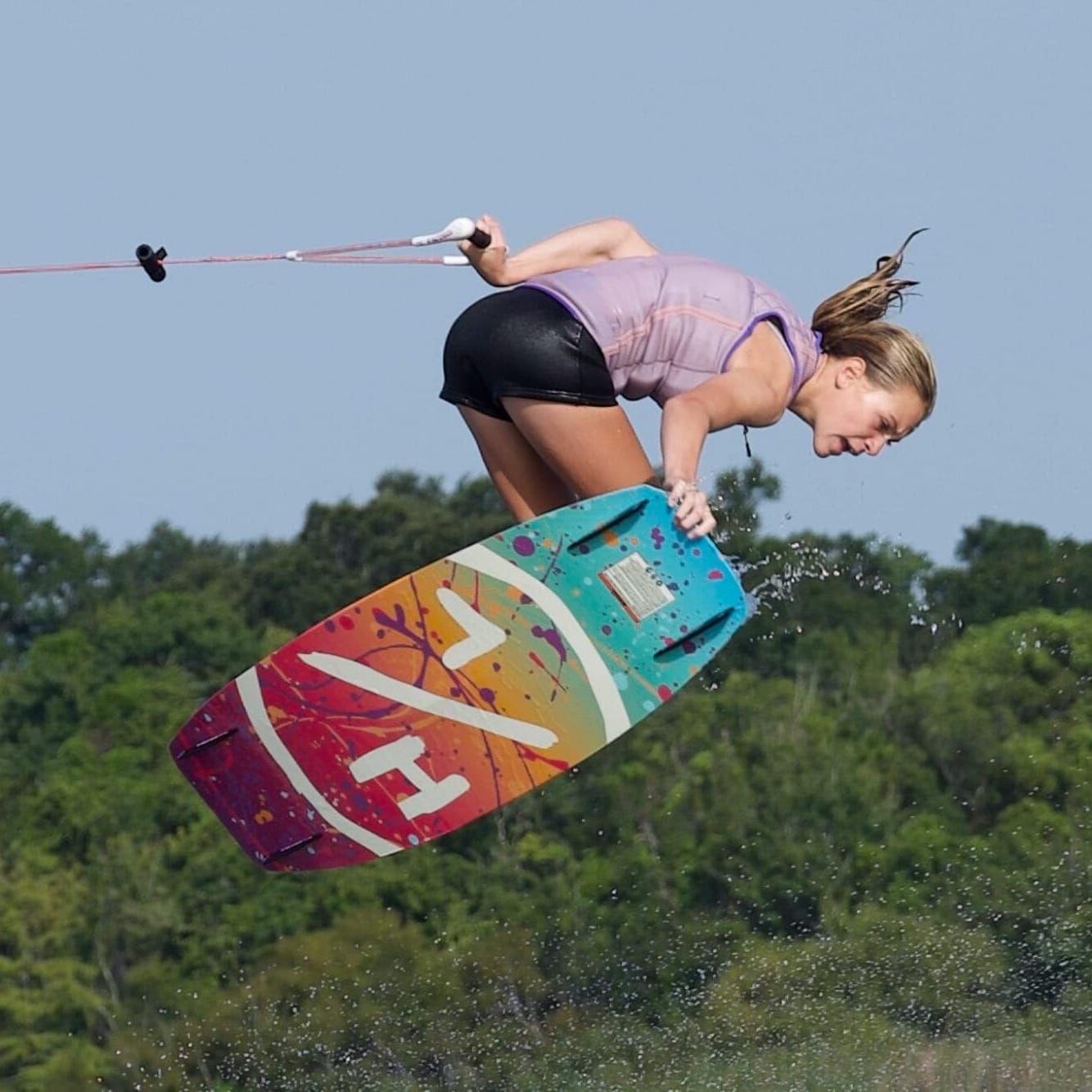 A woman wakeboarding mid-air with a colorful board, holding a tow rope, above a body of water.