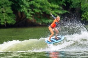 A person wearing an orange life jacket surfs on a river wave, balancing on a blue surfboard with a lush green backdrop.