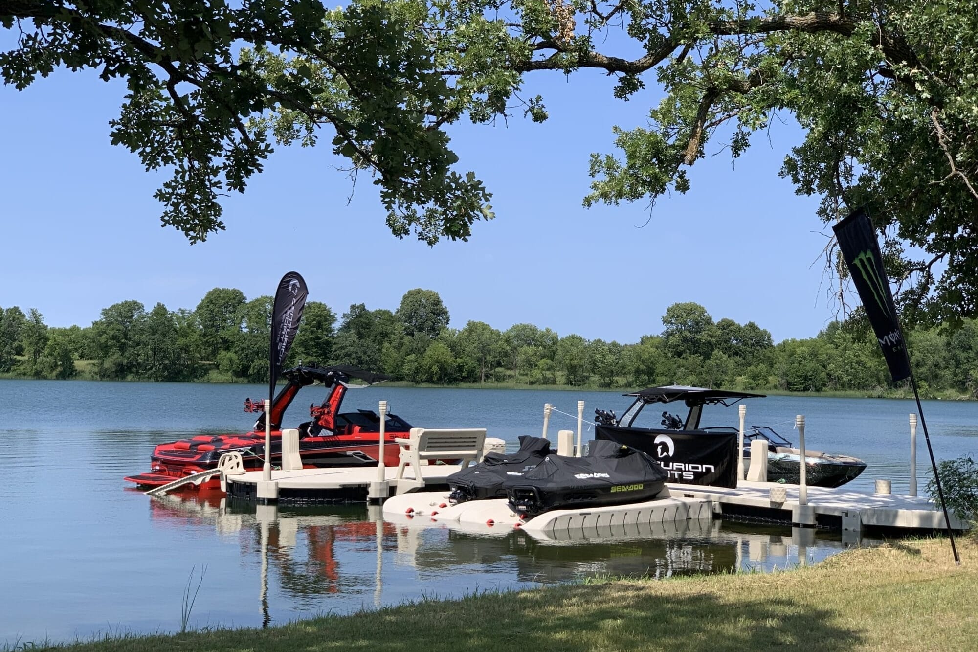 Three boats are docked at a lake shore under a sunny sky. The scene is framed by overhanging tree branches and green grass in the foreground.