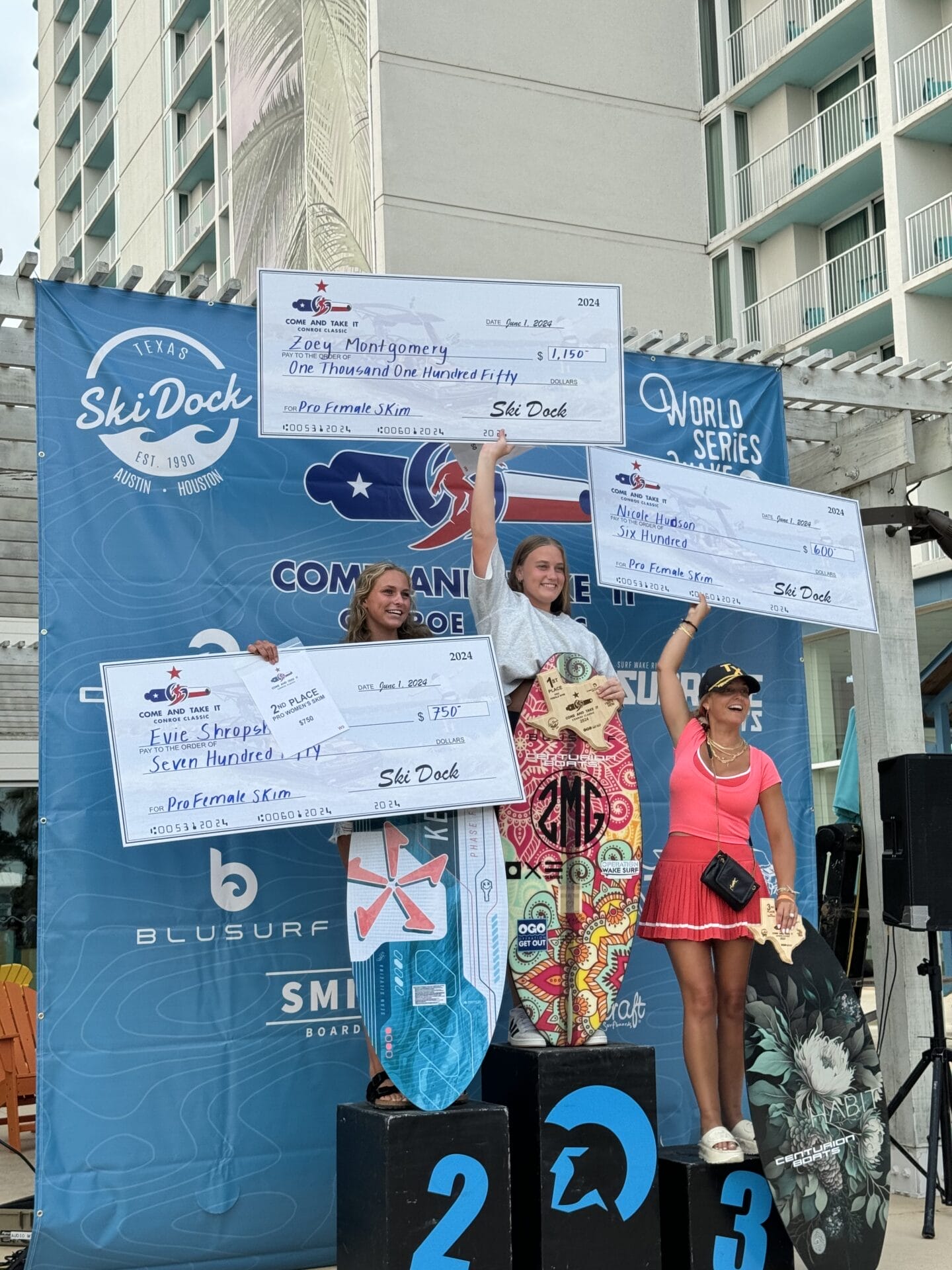 Three female surfers stand on a podium holding large prize checks at a surfing competition. They are smiling, and a surf-themed backdrop displays sponsors' logos.