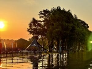 A sunset over a lakeside area with a black pyramid tent labeled "Killjoy Tent" surrounded by trees and a fence, with people visible near the tent and reflections on the water.