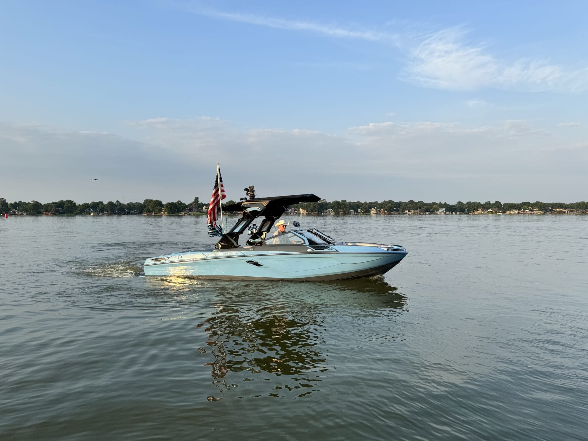 A light blue motorboat with an American flag is moving on a calm lake under a clear sky with some clouds. Trees and houses are visible on the distant shoreline.